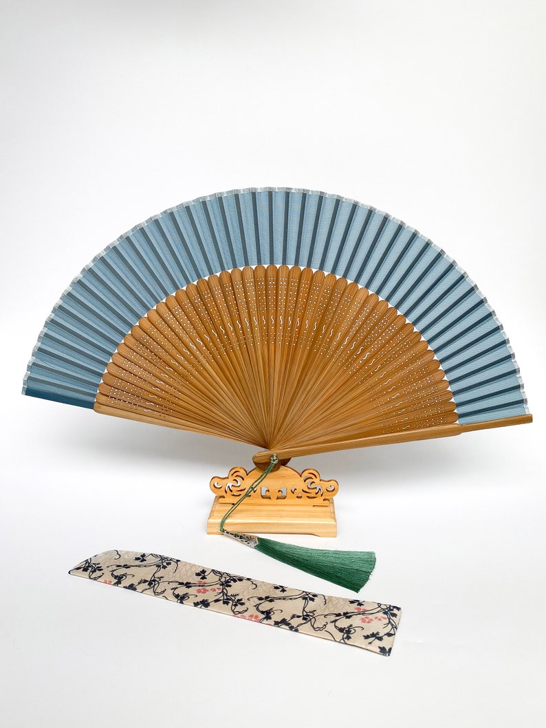 May include: A blue and brown folding fan with a decorative stand and a patterned fabric case. The fan is made of bamboo and has a tassel.