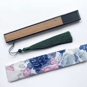 May include: A black and brown folding fan with a green tassel. The fan is closed and has a floral patterned fabric case.