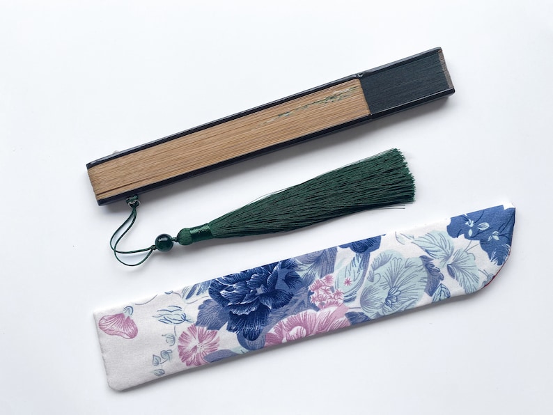 May include: A black and brown folding fan with a green tassel. The fan is closed and has a floral patterned fabric case.