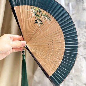 May include: A green and brown folding fan with a floral design and a green tassel. The fan is made of bamboo and has a silk covering.