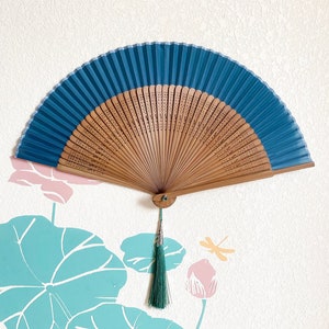 May include: A blue and wooden folding fan with a green tassel hanging from the bottom. The fan is decorated with a floral pattern and is hanging on a white wall.