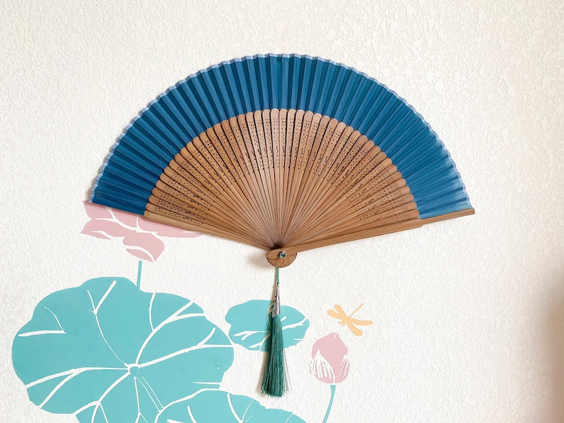 May include: A blue and wooden folding fan with a green tassel hanging from the bottom. The fan is decorated with a floral pattern and is hanging on a white wall.