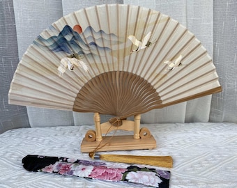May include: A decorative hand fan with a wooden frame and a painted landscape scene featuring mountains, a sun, and cranes. The fan is displayed on a wooden stand, with a matching tassel and a floral-patterned case.