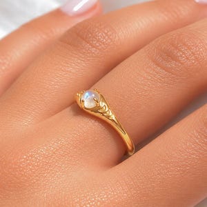 May include: A gold ring featuring a round, iridescent gemstone. The ring has an engraved design on the band and around the gemstone setting. The ring is displayed on a finger, showcasing the jewellery's elegant design.