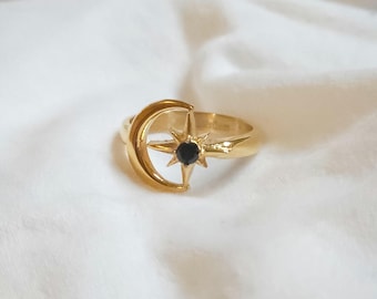 Puede incluir: Un anillo dorado con un diseño de media luna y una estrella con una piedra negra en el centro. El anillo está sobre un fondo blanco suave, resaltando su tema celestial. El diseño del anillo es simple pero elegante.