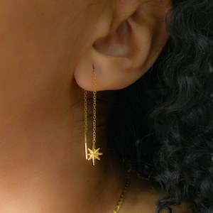 Op de afbeelding: Gouden kettingoorbellen met een stervormige hanger die aan de onderkant hangt. De oorbellen hangen aan de oorlel.
