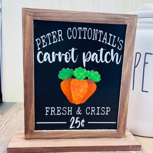 May include: A wooden sign with a chalkboard insert that reads "Peter Cottontail's carrot patch Fresh & Crisp - 25¢". The sign has a brown wooden base and features a painted image of three orange carrots with green tops.