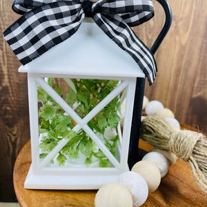 May include: White lantern with a black and white checkered bow, green foliage inside, and a wooden bead garland on a wooden surface.