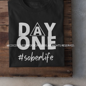 May include: Black t-shirt with white text that reads "DAY ONE" with a triangle containing the letter "A" above the word "ONE". The text "#soberlife" is below the word "ONE". The t-shirt is folded and lying on a wooden surface.