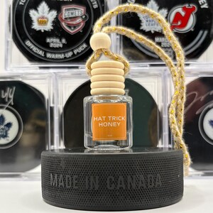 May include: A clear glass bottle with a wooden cap and a gold string hanging from it. The bottle is labelled "Hat Trick Honey". The bottle is sitting on a black hockey puck with the words "Made in Canada" printed on it.