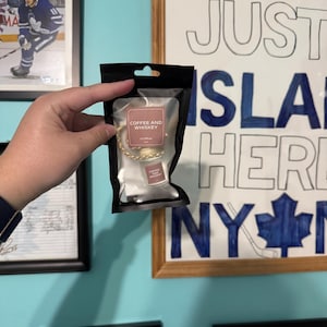 Op de afbeelding: Een auto luchtverfrisser in een zwart pakket. Het label leest "COFFEE AND WHISKEY". De luchtverfrisser is een kleine, rechthoekige vorm met een goudkleurige ketting. De achtergrond bevat ingelijste foto's en een bord met "JUST ISLAND HERE NY".