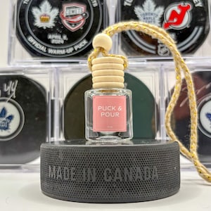 May include: A clear glass bottle with a pink label that reads "PUCK & POUR" is suspended by a gold string. The bottle sits on a black hockey puck with "MADE IN CANADA" imprinted. Hockey pucks with team logos are in the background.
