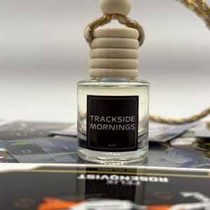 May include: A clear glass bottle with a wooden cap and a gold string. The bottle is labeled "TRACKSIDE MORNINGS" and contains 8 ml of liquid.