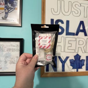 May include: A black package containing a small car air freshener with a candy cane scent. The label reads "CANDY CANE" in red and white. The package is held in front of a blue wall with framed pictures and a sign that says "JUST ISLAND HERE NY".