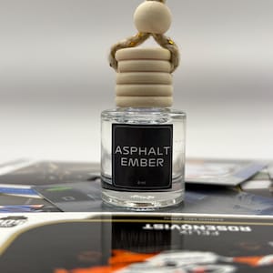 Op de afbeelding: Een heldere glazen fles met een zwart etiket waarop "Asphalt Ember" staat en een houten dop. De fles hangt aan een gouden en witte touwtje.
