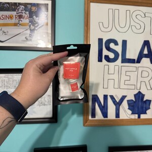 May include: A black pouch with a clear window containing a small, red label that reads "HOT APPLE PIE". The pouch is held in front of a light blue wall with framed pictures and a drawing that says "JUST ISLAND HERO NY".