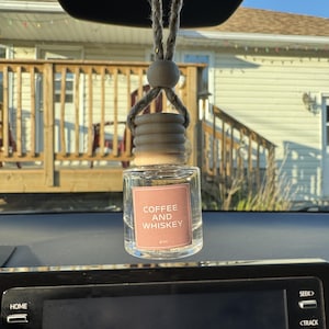 May include: A small, clear glass car air freshener bottle with a light wooden cap and a jute rope for hanging. The bottle features a square pink label with the white text COFFEE AND WHISKEY and 8ml printed on the front, suspended by a loop.
