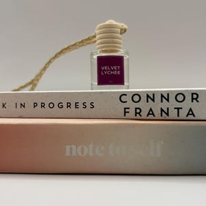May include: A small, clear glass bottle with a wooden cap and a gold string, labeled "VELVET LYCHEE" in white text on a magenta background. The bottle sits atop two books, one reading "CONNOR FRANTA" and the other "note to self".
