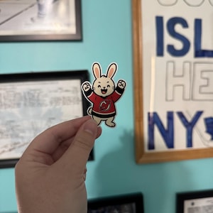 May include: A sticker featuring a cartoon bunny wearing a red and black hockey jersey with the letter "C" on the chest. The bunny has its arms raised in a celebratory gesture. The background includes framed pictures and a sign that says "ISL HE NY".