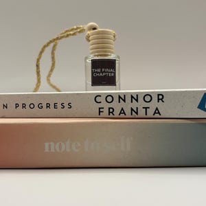 May include: A small, clear glass bottle with a wooden cap and a gold string, labelled "THE FINAL CHAPTER." The bottle sits atop two books, one titled "CONNOR FRANTA" and the other "note to self."