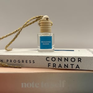 May include: A small, clear glass bottle with a wooden cap and a blue label reading "READING NOOK." The bottle hangs by a gold-flecked string and sits on a book titled "CONNOR FRANTA." Another book, "note to self," is visible below.