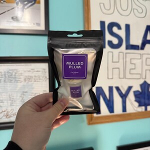 May include: A black pouch with a silver interior, holding a car air freshener. The label reads "MULLED PLUM" in white text on a purple background. The pouch is held in front of a wall with framed pictures and a sign that says "JUST ISLAND HER NY".