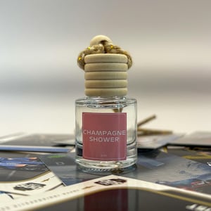 May include: A small glass bottle with a wooden top and a pink label that says "Champagne Shower" with a gold string attached to the top. The bottle is sitting on a table with other items.