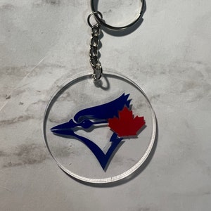 May include: A clear acrylic keychain with a blue and white Toronto Blue Jays logo. The logo features a stylized blue jay with a red maple leaf on its wing.