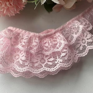 May include: Close-up of a delicate, light pink lace trim with a ruffled edge. The trim features a layered design, with intricate floral patterns visible throughout the lace. The soft pink color and delicate texture suggest its use in crafting or garment embellishment.