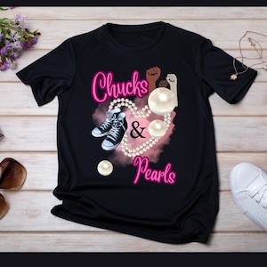 Puede incluir: Camiseta negra con las palabras "Chucks & Pearls" en escritura rosa. El diseño incluye un collar de perlas en forma de corazón, dos zapatillas deportivas en blanco y negro y detalles de perlas. La camiseta también presenta dos puños levantados, uno marrón y otro blanco.