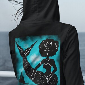 Könnte beinhalten: Schwarzer Kapuzenpullover mit einem schwarz-weißen Meerjungfrau-Motiv auf dem Rücken. Die Meerjungfrau trägt eine Krone und Halskette, vor einem türkisfarbenen und blauen Hintergrund. Der Hoodie hat eine Kapuze und lange Ärmel.