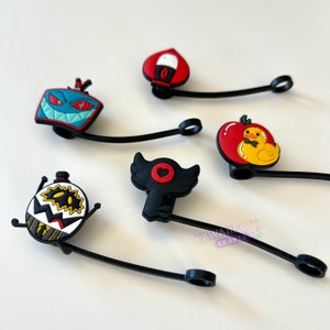 Peut inclure: Six bouchons de bouteille en silicone colorés avec des pailles flexibles noires. Les bouchons présentent divers personnages de dessins animés, notamment un cœur rouge avec des ailes, une pomme rouge avec un canard jaune, un visage de monstre bleu et vert et un monstre noir et blanc avec un chapeau haut de forme.