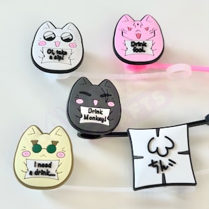 May include: Four cat-shaped phone charms with different expressions and messages. The charms are made of silicone and have a black, white, pink, and yellow color scheme. The messages on the charms read "Drink Fool", "Ol, take a sip", "Drink Monkey", and "I need a drink".