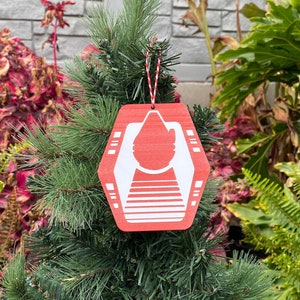 May include: A red and white Christmas ornament shaped like a hexagon with a white symbol inside. The ornament is hanging on a small artificial Christmas tree.
