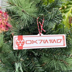 May include: A red and white Christmas ornament with a white background and red text that reads "OK7U1W1D". The ornament is hanging on a green Christmas tree.
