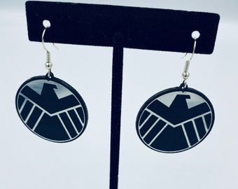 May include: A pair of black and silver earrings featuring the logo of S.H.I.E.L.D., a fictional organization from the Marvel Universe.