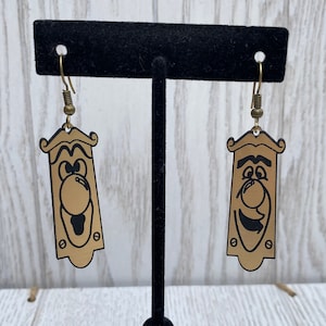 May include: A pair of gold-colored door knob earrings with black cartoon faces. The earrings are hanging from a black earring stand.