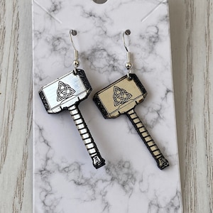 May include: A pair of silver earrings shaped like Thor's hammer with a black and gold design and a Celtic knot on the hammer head.