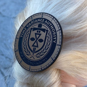 May include: A round, brown leather pin with a black and gold design. The pin features a sword, a shield with a stylized 'X' inside, and a wreath of leaves. The text around the design reads "Time Variance Authority" and "For All Time, Always".