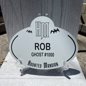 May include: White oval name tag with black text. The text reads "HM", "ROB", "GHOST #1000", and "HAUNTED MANSION". There are two black bats on either side of the "HM".