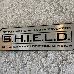 May include: A silver metal sign with black text that reads "Strategic Homeland Intervention, S.H.I.E.L.D., Enforcement Logistics Division".