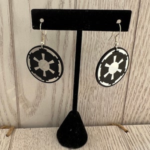 May include: Black and silver earrings featuring the Galactic Empire symbol. The earrings are circular with a black background and a silver emblem. They hang from silver fish hook ear wires and are displayed on a black earring stand.