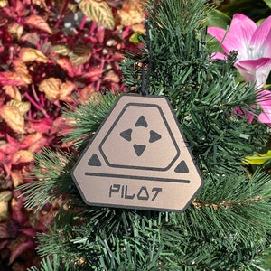 May include: A brown and black ornament shaped like a triangle with rounded edges. The ornament has a black outline and four black arrows pointing inward. The word "PILOT" is printed in black letters on the ornament.