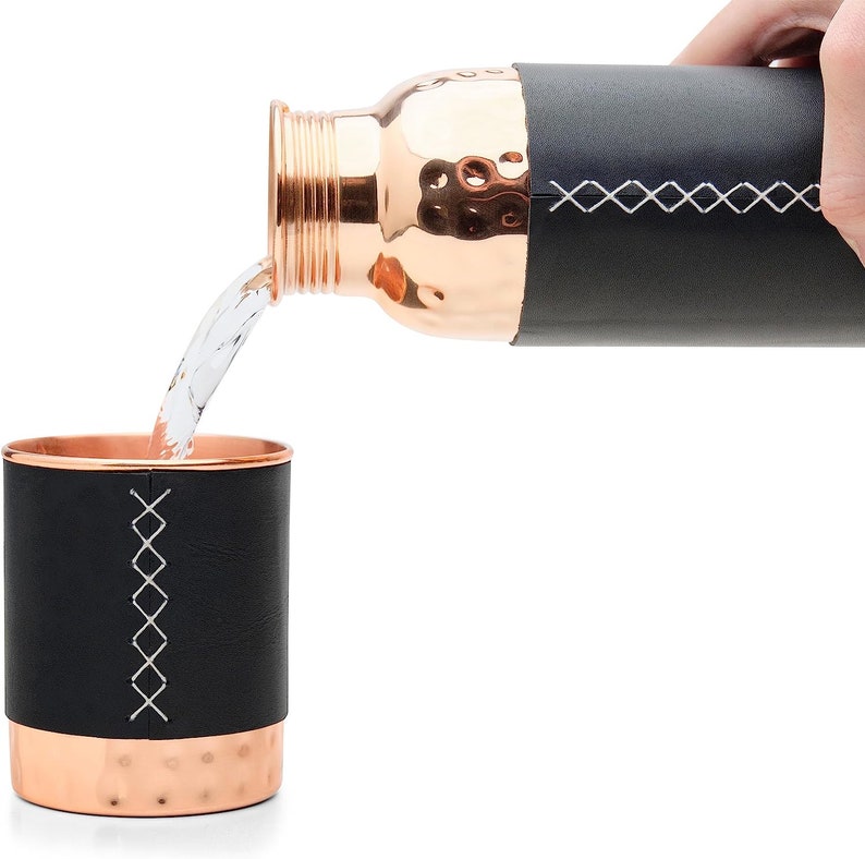 Puede incluir: Una botella de agua de cobre con una funda de cuero negro y un dise&ntilde;o cosido est&aacute; vertiendo agua en una taza de cobre con una funda de cuero negro y un dise&ntilde;o cosido.