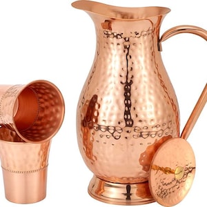 May include: A set of three copper drinking vessels, including a pitcher and two tumblers. The pitcher has a hammered finish and a wide, rounded spout. The tumblers are also hammered and have a slightly flared lip.