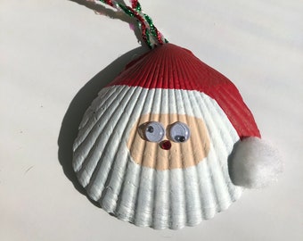 May include: A white seashell painted to look like Santa Claus with a red hat, white beard, and googly eyes. The seashell is attached to a red, green, and white string.