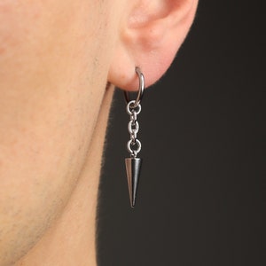 May include: A silver-colored earring with a chain and a cone-shaped pendant. The earring is attached to a hoop that is fastened to the ear. The earring is shown against a dark background.