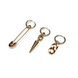 May include: Three gold-colored earrings on a white background. One earring is a safety pin, another has a cone-shaped charm, and the third has a chain link design. The earrings are likely made of metal.