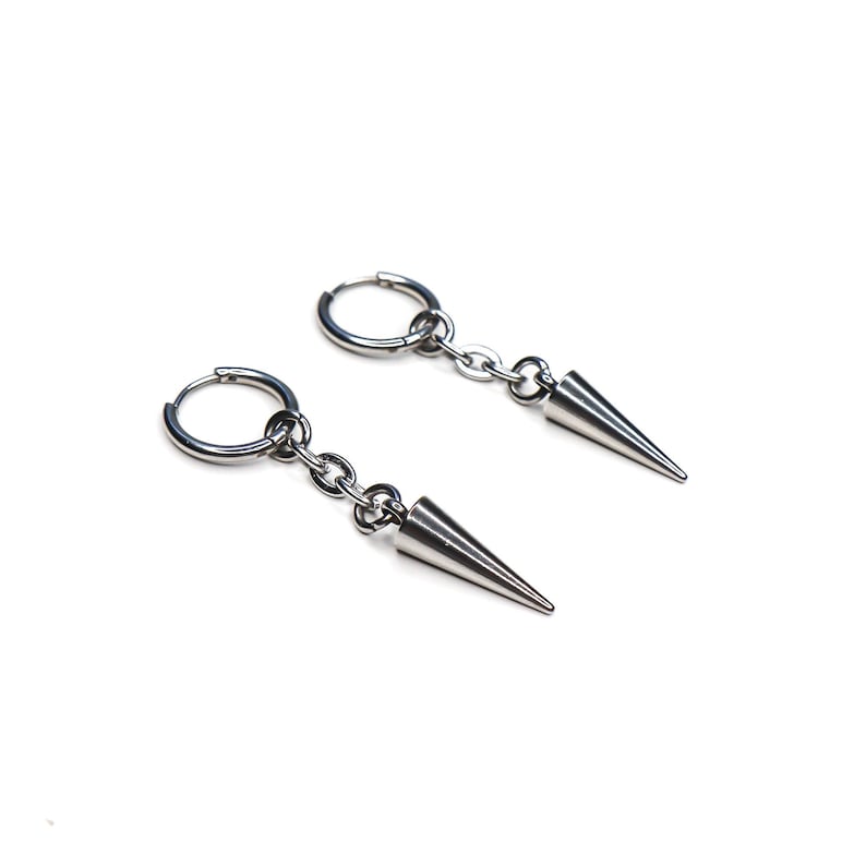 May include: A pair of silver-colored hoop earrings with a chain and a conical spike pendant. The earrings are displayed on a white background, showcasing their metallic sheen and minimalist design. The jewelry is suitable for various styles.