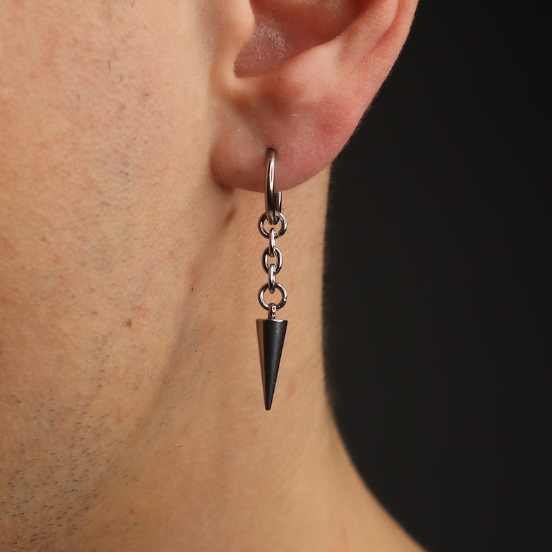May include: A silver-toned earring with a conical pendant. The earring features a small hoop that connects to a series of small chain links, which then connect to the dark gray, cone-shaped pendant. The earring is shown against a dark background.